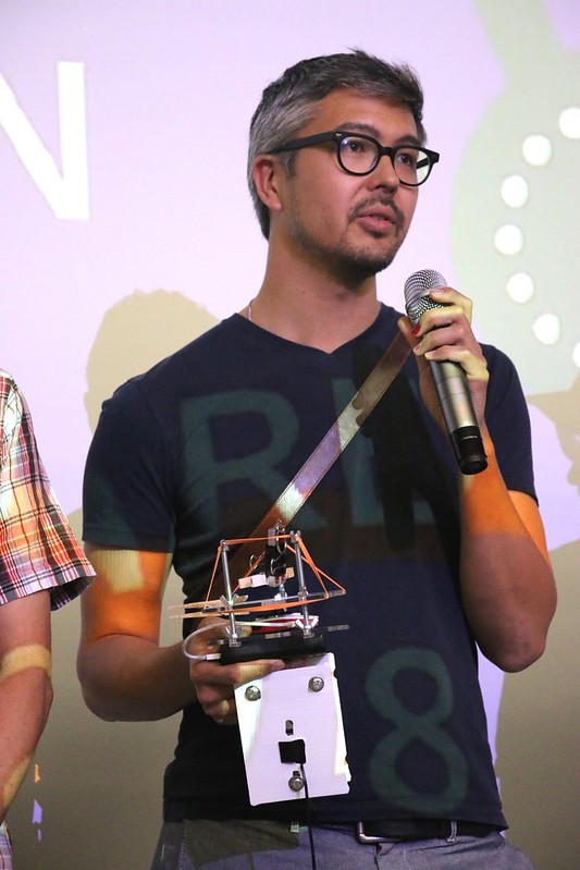 Photo of attendee holding a piece of hardware while speaking into a microphone.